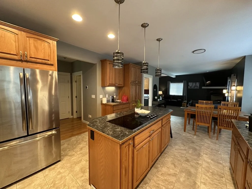 Modern kitchen with stainless steel refrigerator, wooden cabinetry, and a black countertop island. There are pendant lights hanging above the island and a dining area with wooden chairs in the background.