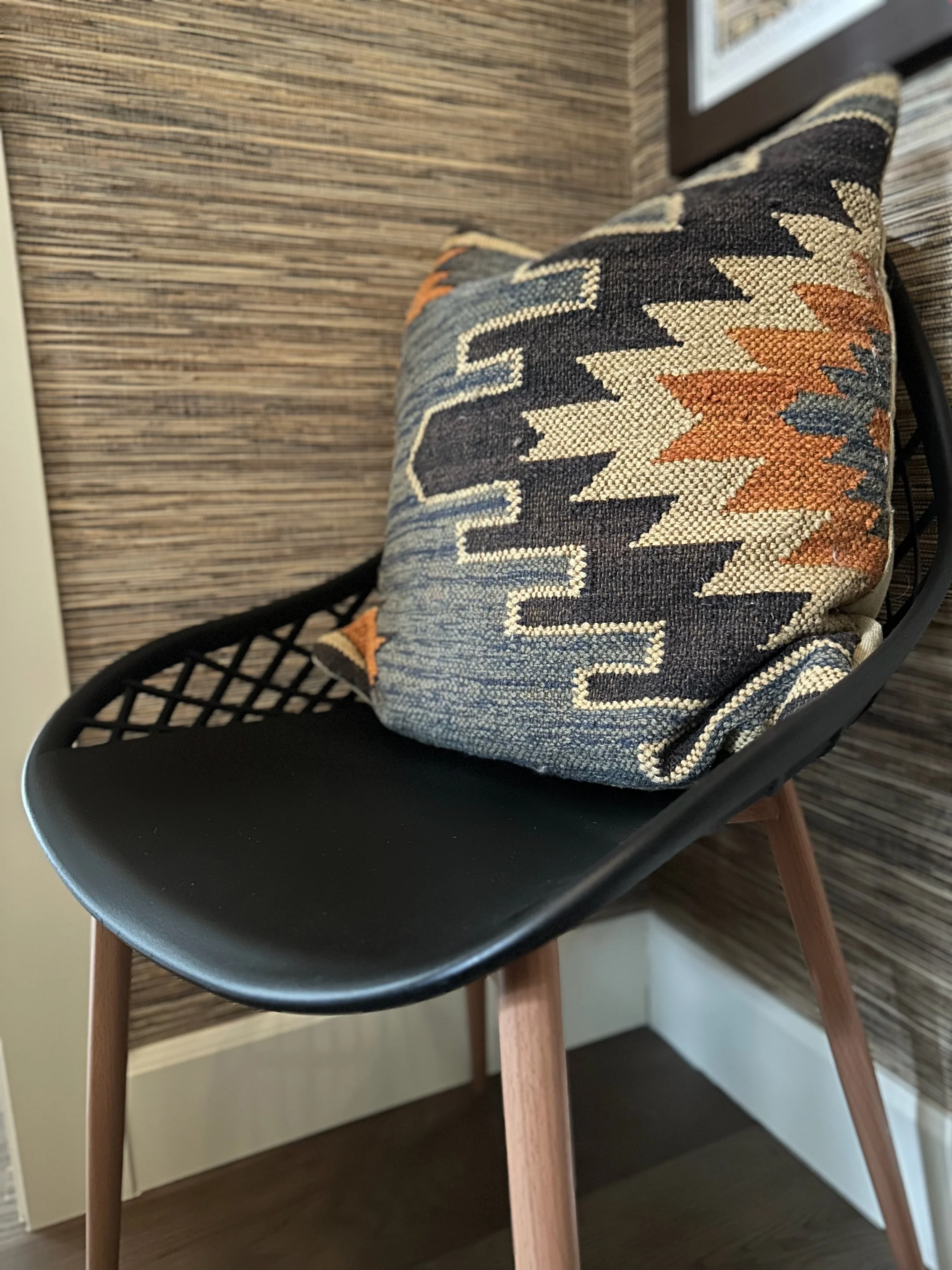 Black chair with wooden legs and a woven geometric-patterned pillow in a corner with textured wallpaper.