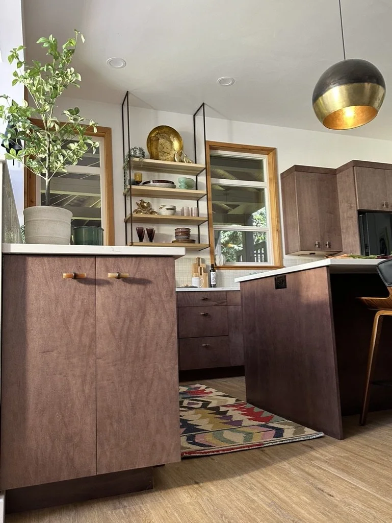 Modern kitchen interior with wooden cabinets, open shelving, a hanging light fixture, and a colorful geometric rug. A potted plant is on the countertop, and decorative items are on the shelves.