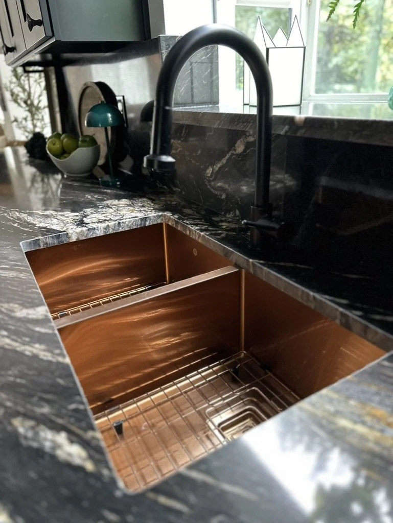 Modern kitchen sink with black countertop, copper double basin, and a black faucet. In the background, a bowl with green apples and a small teal lamp are visible on the counter.