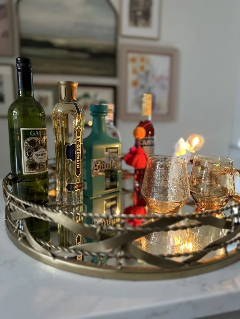 A decorative tray with various liquor bottles and two glasses on a countertop with framed artwork in the background.