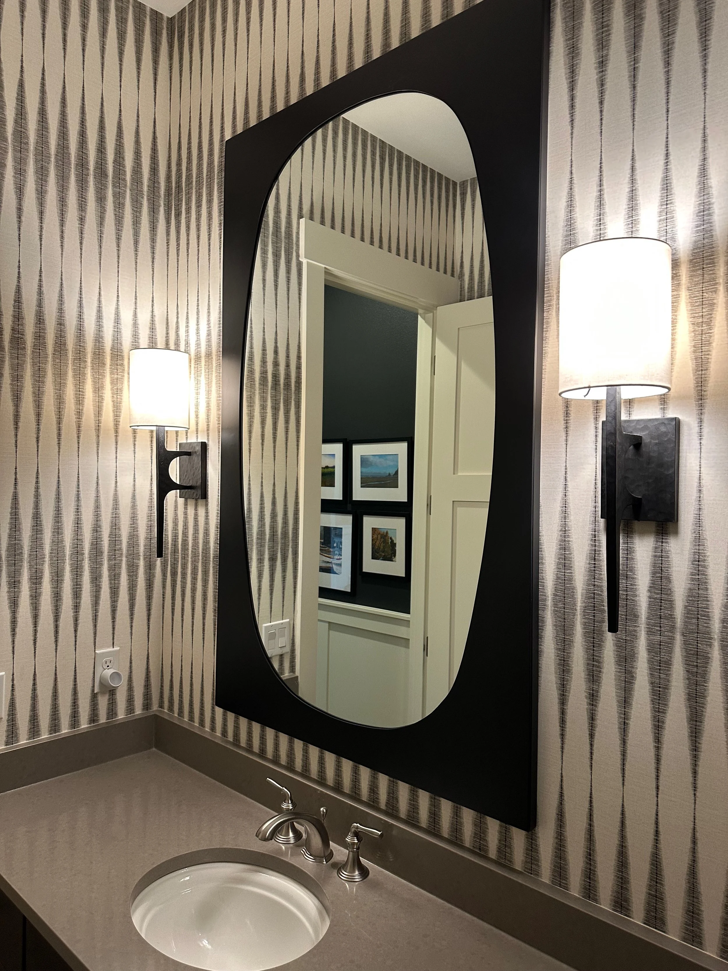 Modern bathroom with geometric wallpaper, a large mirror, dual wall sconces, a sink with silver faucet, and framed pictures in the background.