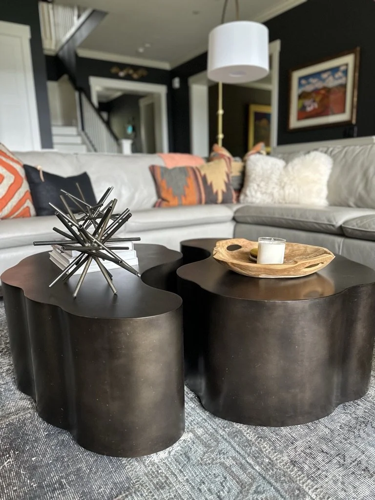 Modern living room with black abstract-shaped coffee tables, a white candle on a wooden tray, decorative metal sculpture, gray sofa with colorful pillows, black walls, and artwork.