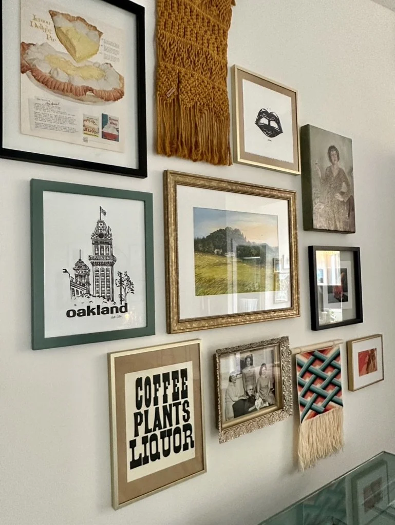 A gallery wall featuring a mix of framed artworks, including a yellow fringe macrame, a vintage pie advertisement, an illustration labeled 'Oakland,' photography, typography art stating 'COFFEE PLANTS LIQUOR,' and other assorted prints and photograph