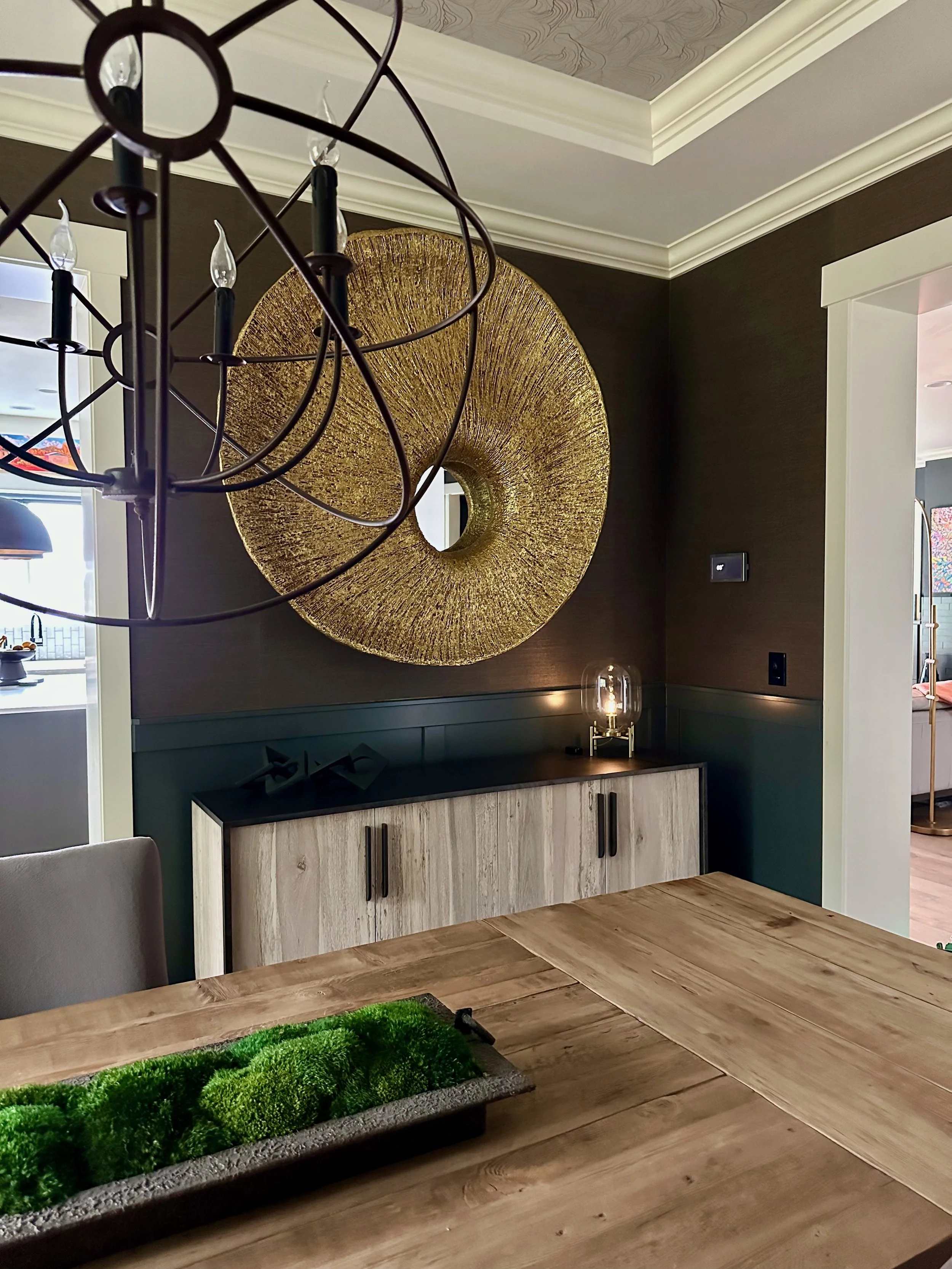 Modern dining room with a large circular gold wall mirror, wood table, green decorative centerpiece, and black metal chandelier.