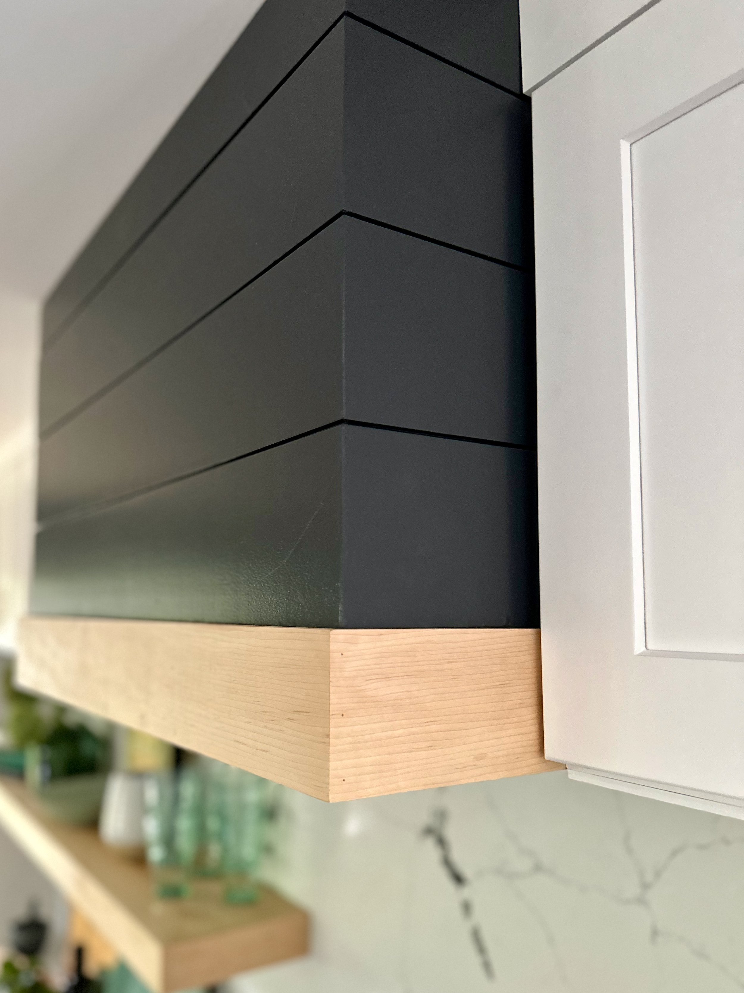 Modern kitchen with minimalist black and wood range hood, white cabinets, and open shelves with glassware and decor items.