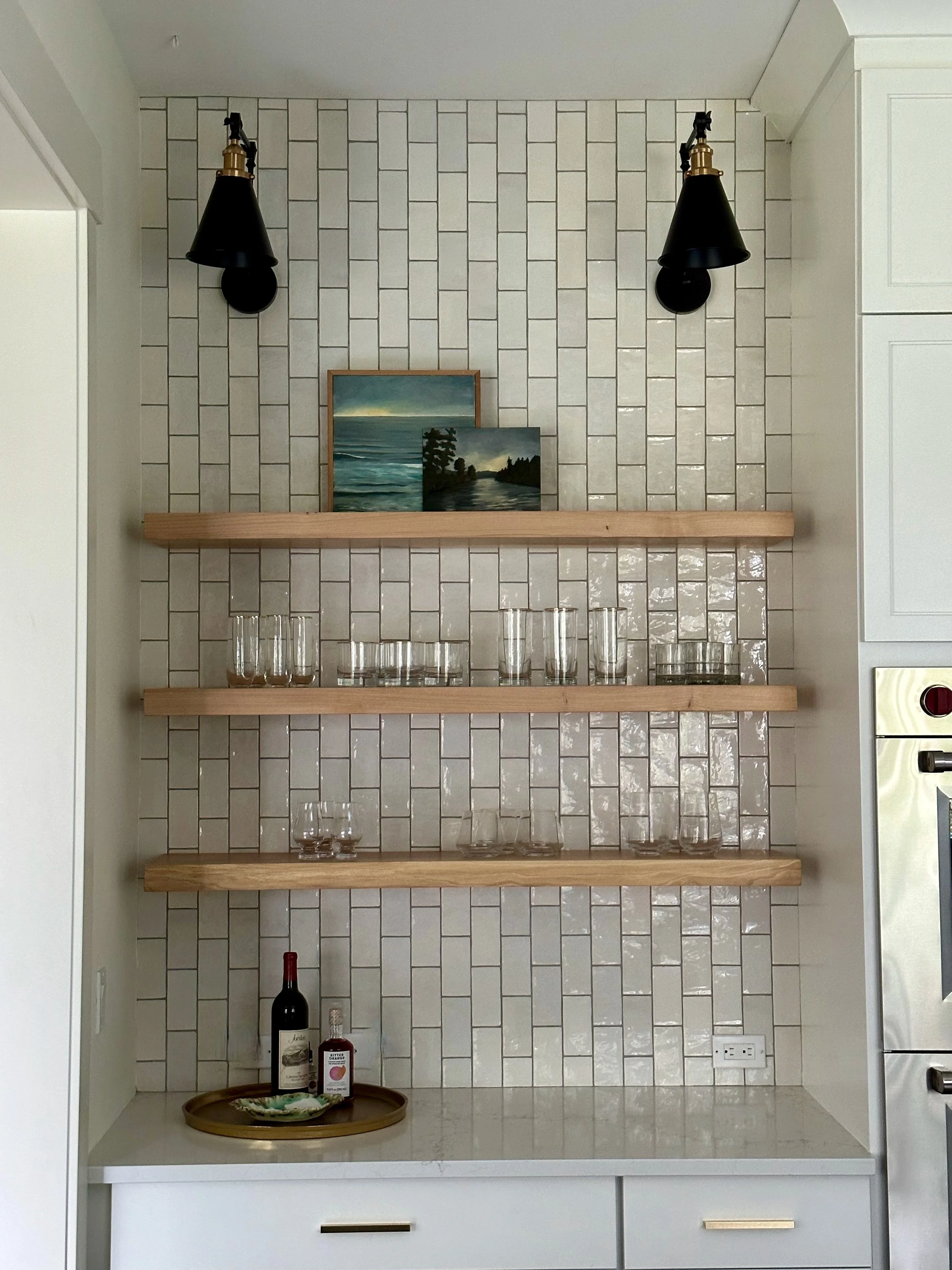 Kitchen nook with white subway tile wall, wooden shelves holding glasses, a painting, and a tray with wine and bitters on a countertop.