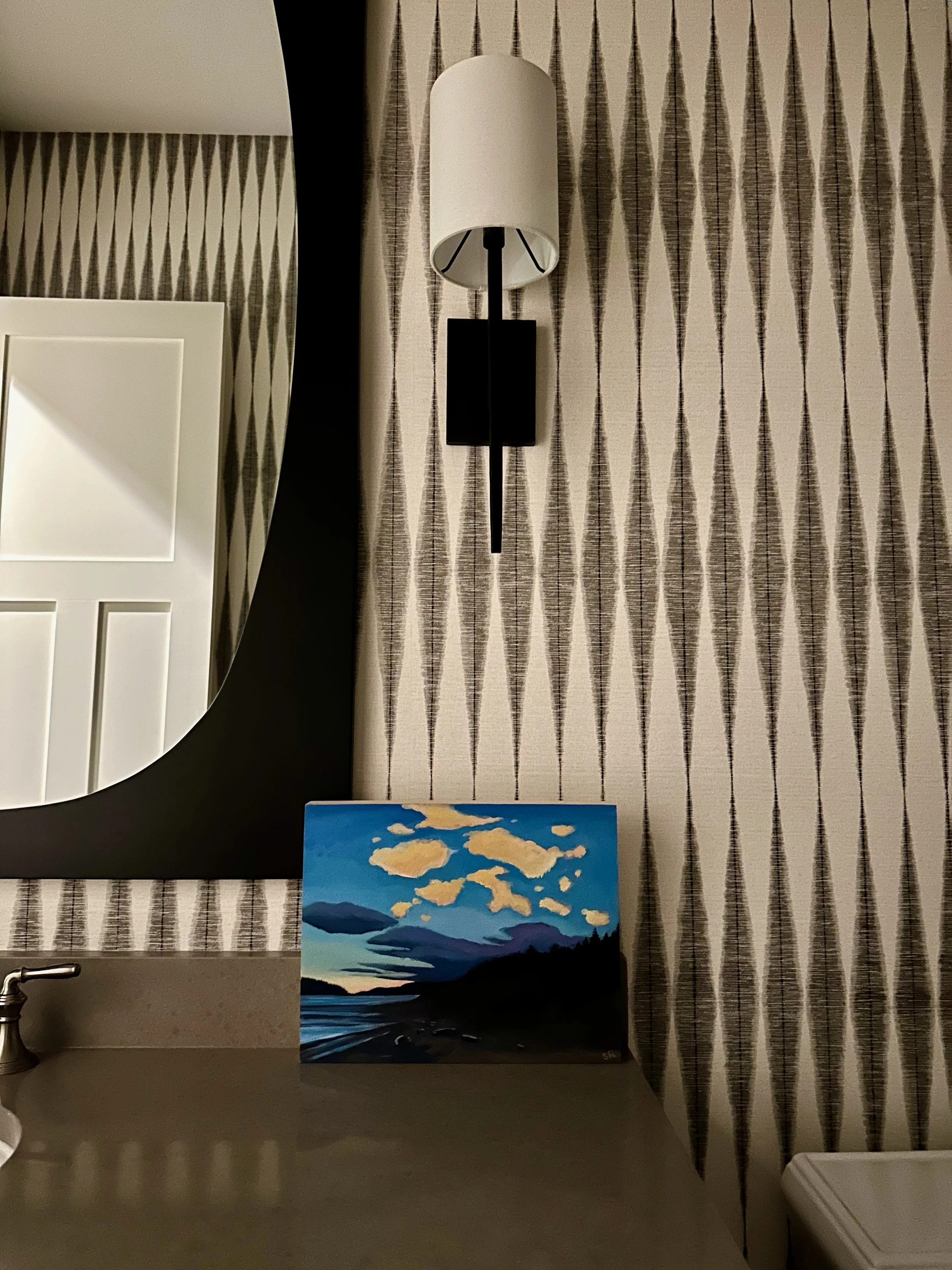 Bathroom with geometric patterned wallpaper, a wall-mounted light fixture, a dark-framed mirror, a countertop with a small landscape painting, and a sink faucet visible.