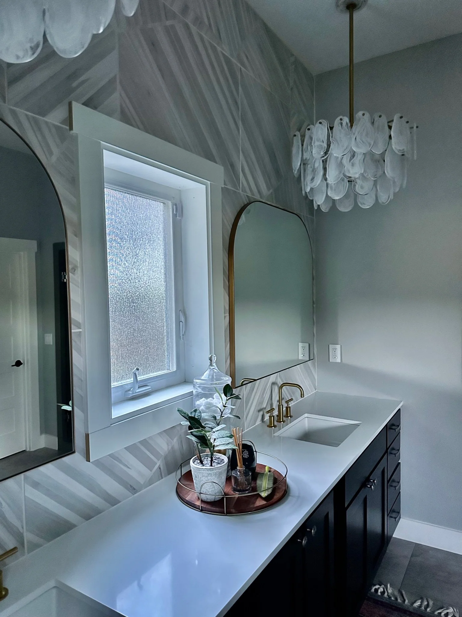 Elegant bathroom with a double vanity, white countertops, gold faucets, large mirrors, a decorative plant, a chandelier, and a frosted window.