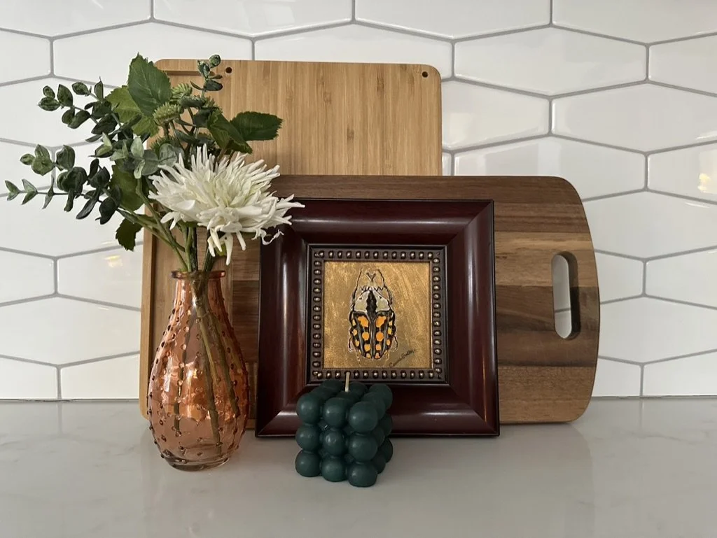 Tabletop with decorative arrangement including a vase with white flowers and greenery, a framed beetle artwork, two wooden cutting boards, and a teal bubble candle against a tile backsplash.