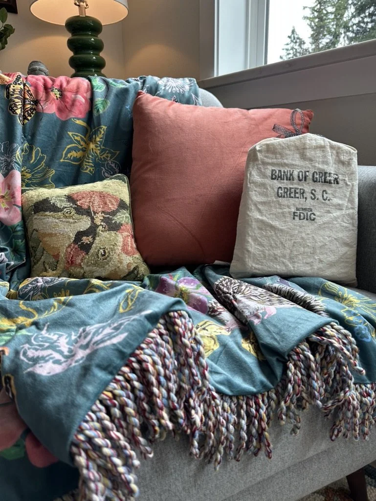 A cozy living room setup with a floral blanket, a pink pillow, a decorative pillow, and a retro-style fabric bag labeled with "Bank of Greer, Greer, S.C., FDIC" on a sofa. A green lamp and a window are in the background.
