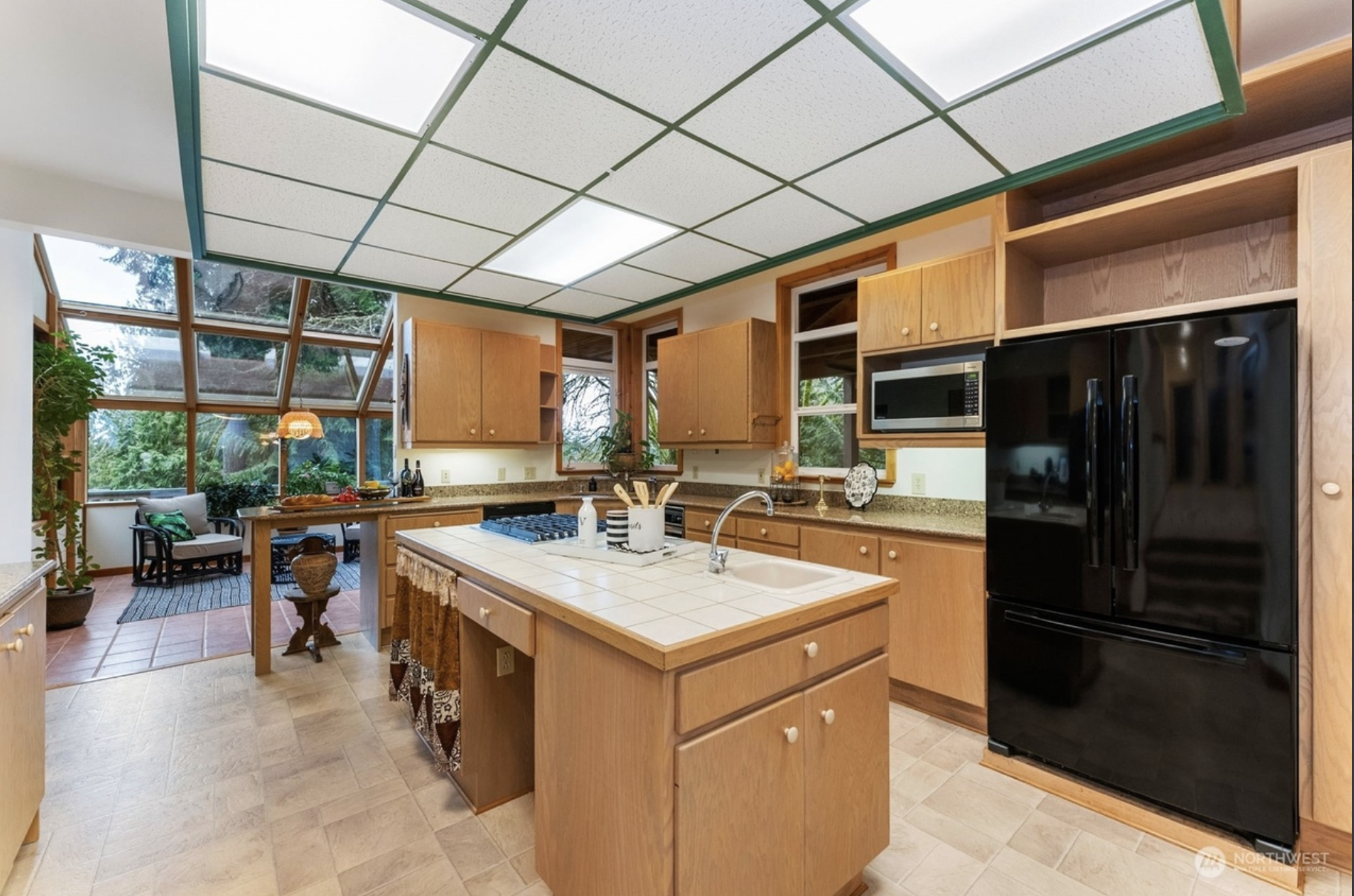 Modern kitchen with wooden cabinets, black refrigerator, tile countertops, kitchen island with sink, and large windows with outdoor view.