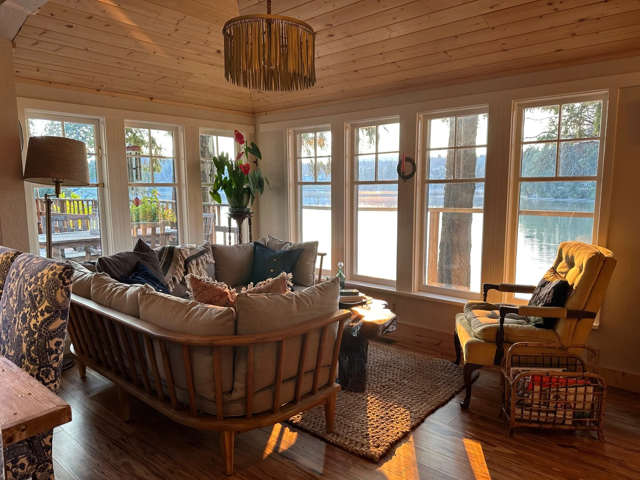Cozy living room with wooden ceiling and large windows overlooking a lake. Features a wicker couch with cushions, a plant, a yellow armchair, and a wicker basket on a rug.