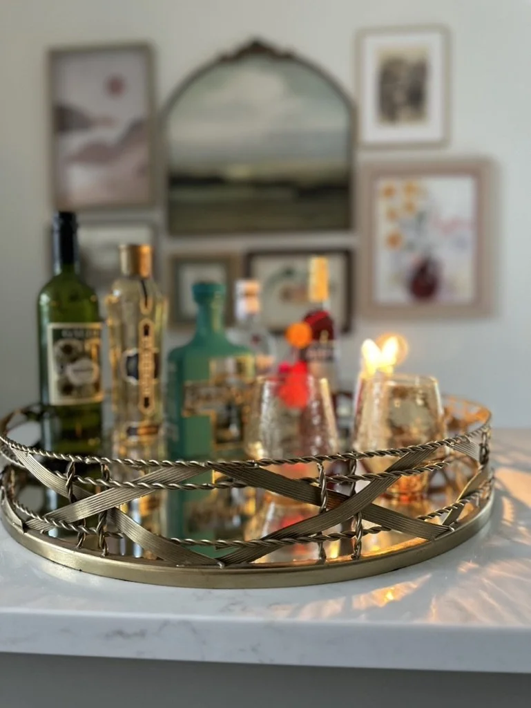 Decorative tray with liquor bottles and glasses on a marble surface, with framed artwork in the background.