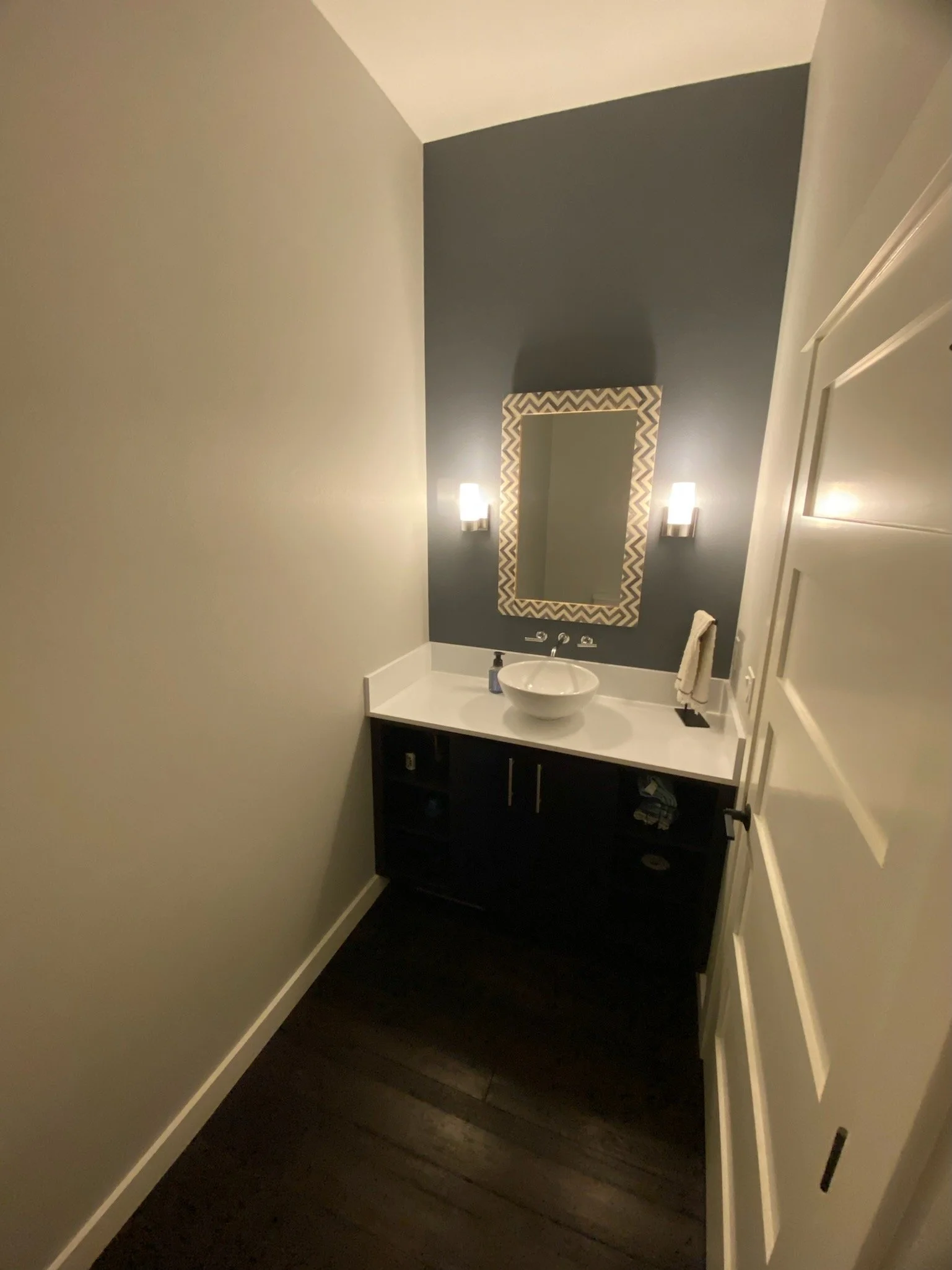 Modern bathroom with a dark accent wall, white porcelain sink, rectangular mirror, wall-mounted lights, and wooden floor.