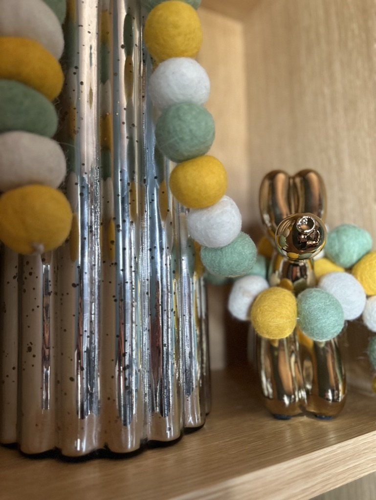 Close-up of decorative glass object with colorful felt ball garland and small gold-colored bunny figurine on a wooden shelf.