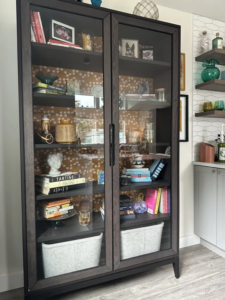 A black wooden cabinet with glass doors, containing books, decorative items, and photos. The background features a patterned design. There are two storage bins at the bottom shelf and various items like glassware and a bust are displayed on the shelv