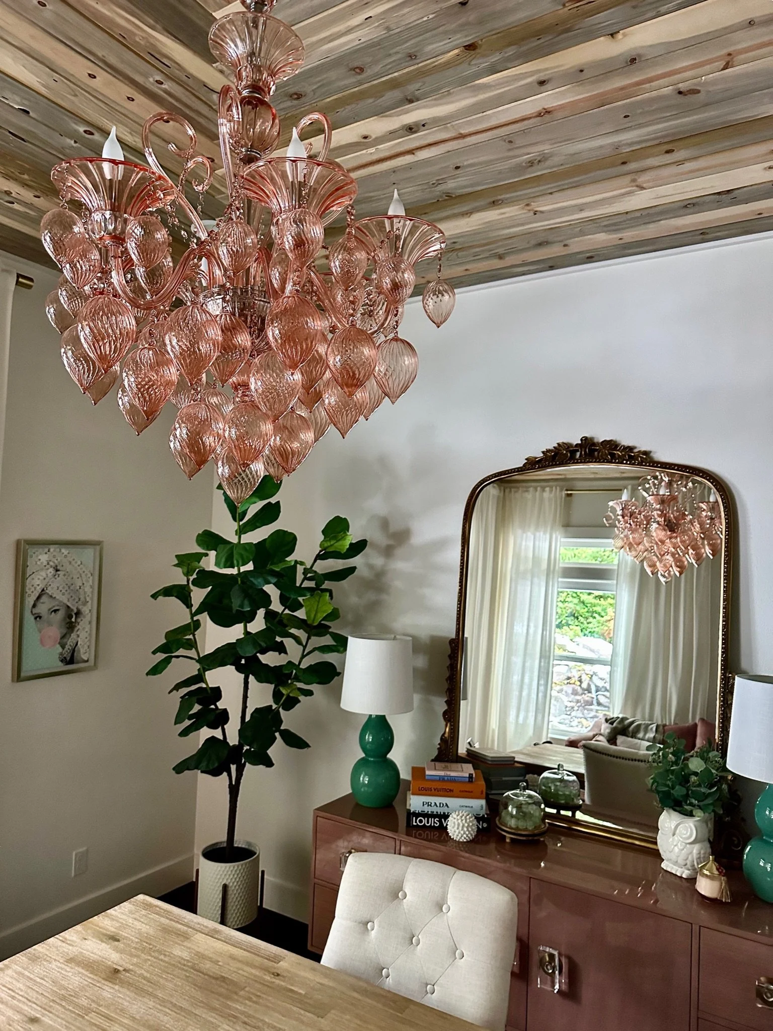Interior room with a large pink glass chandelier hanging from a wooden ceiling, a wooden desk with a beige chair, a green ceramic lamp, potted plant, wall art, and a dresser with books and decorations including a mirror reflecting a window view.