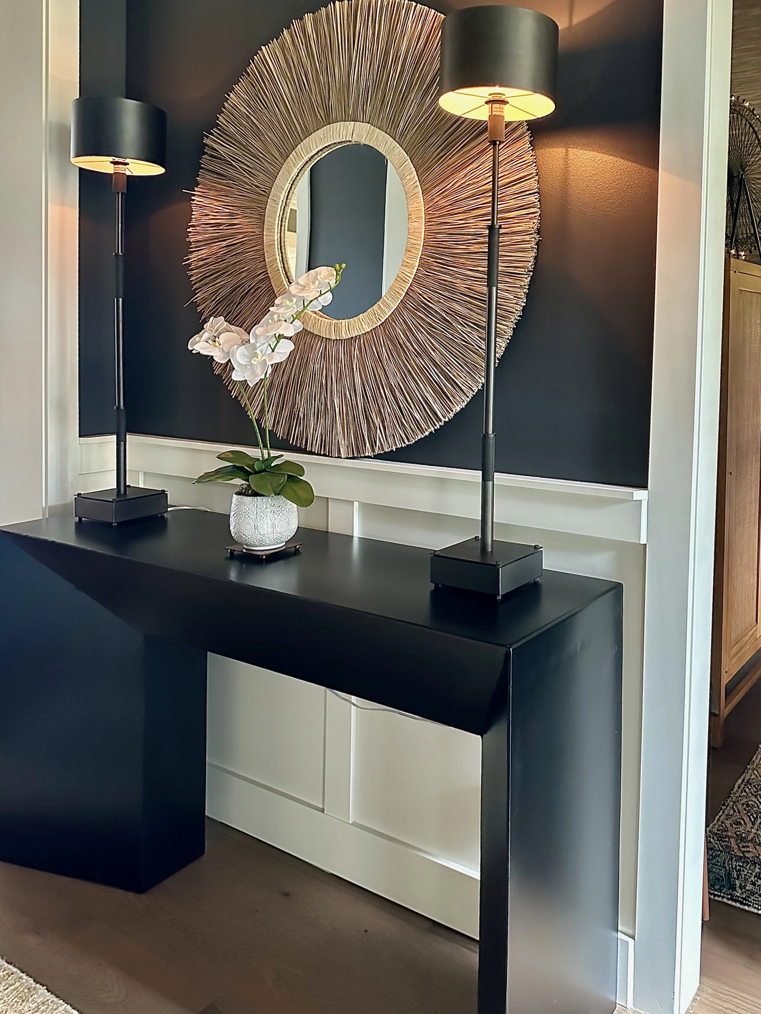 A black console table with two modern black lamps and a white orchid in a vase, positioned against a dark wall. A large round decorative mirror with a textured sunburst frame hangs above the table.