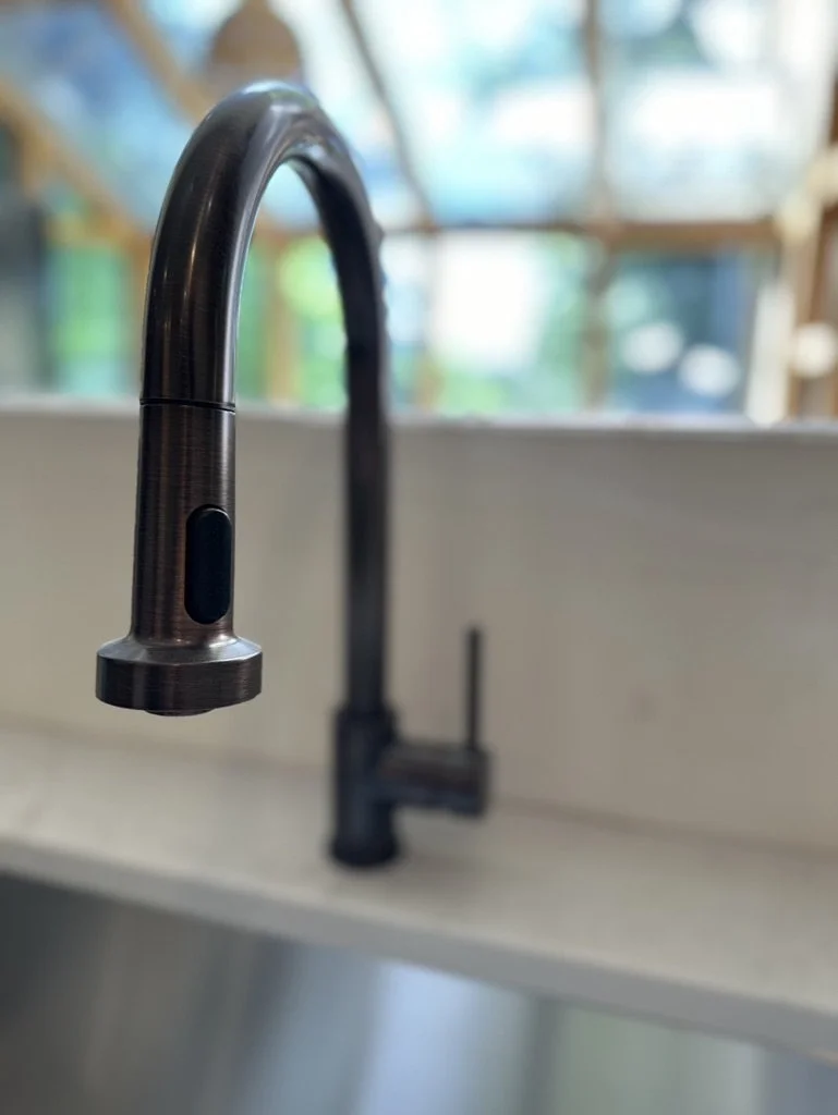 Close-up of a modern kitchen faucet with a gooseneck design and spray nozzle, against a blurred background of a kitchen window.
