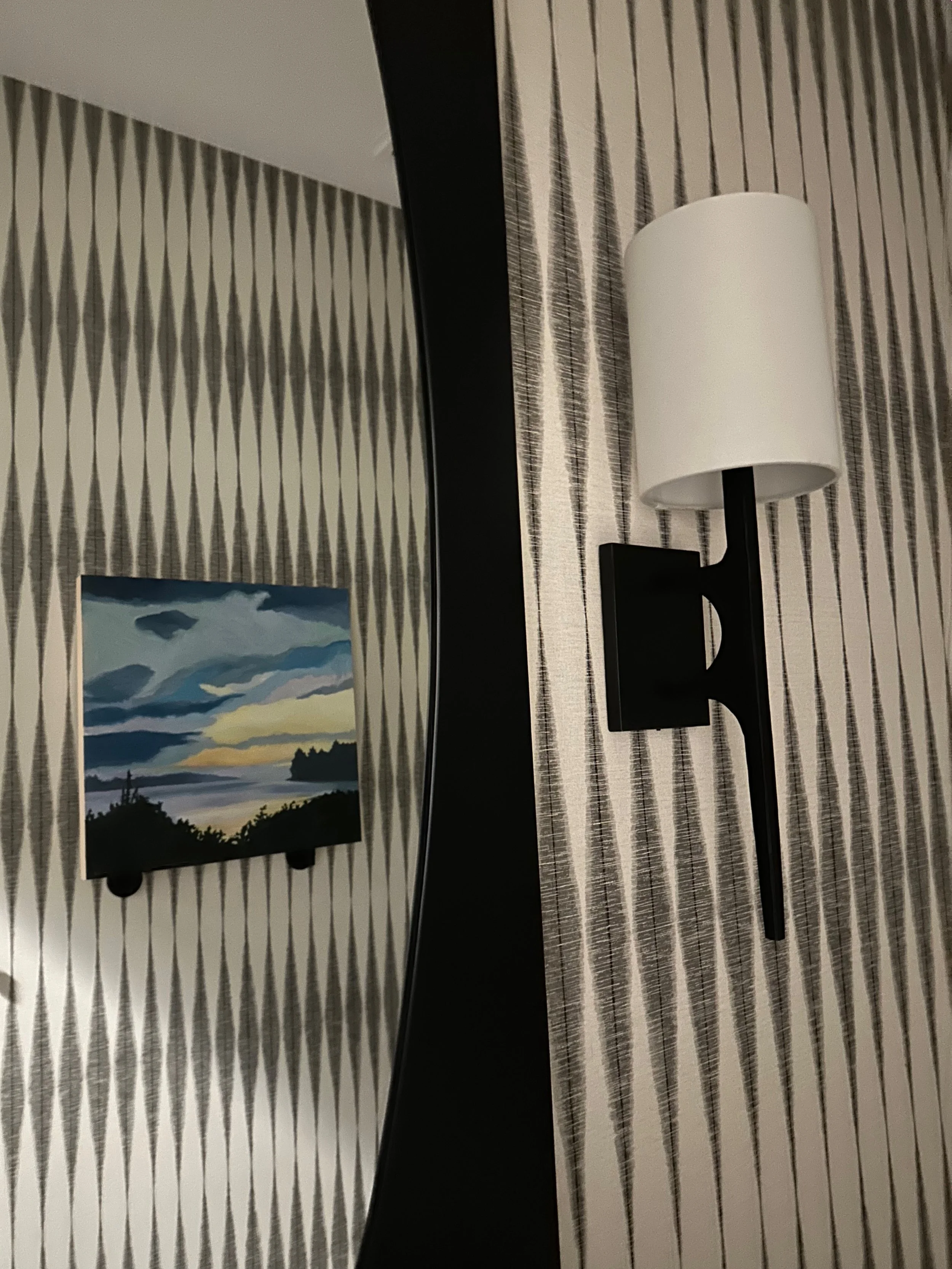 Interior wall with geometric wallpaper, featuring a black wall sconce with a white cylindrical shade and a small framed painting of a landscape reflecting in a mirror.