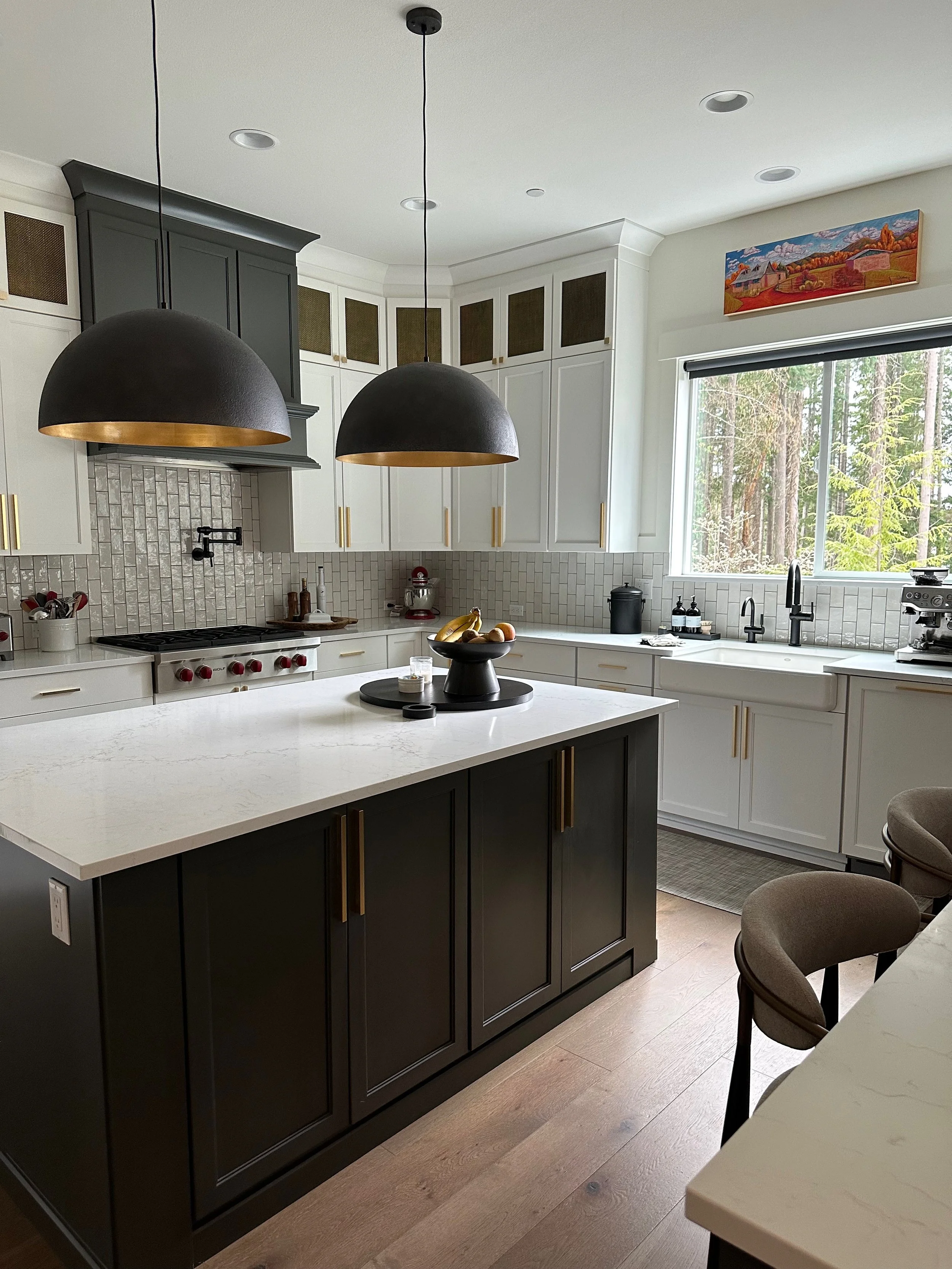 Modern kitchen interior with white and dark green cabinetry, white marble countertops, large pendant lights, and window view. Island with decorative bowl and bar stools. Stainless steel appliances and accessories.