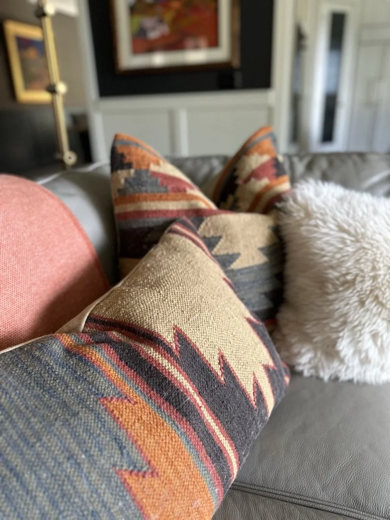 Close-up of decorative pillows on a leather couch, featuring a geometric pattern in earthy tones and a fluffy white accent pillow. Blurred background includes wall art and part of a lamp.