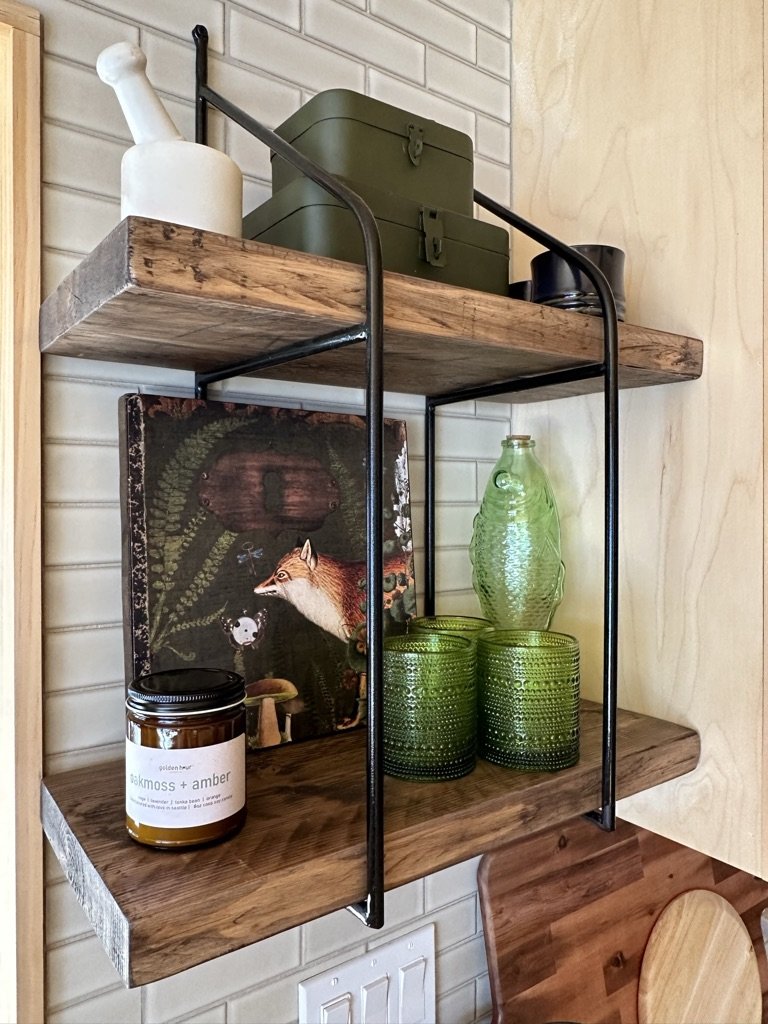 Wooden shelves with a green metal storage box, a white mortar, a decorative fox art piece, a scented candle labeled 'oakmoss + amber,' a green glass bottle, and four green textured glasses.