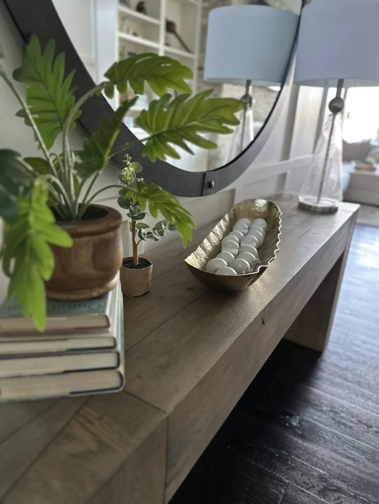 Wooden console table with houseplants, a stack of books, decorative bowl with white orbs, and a round mirror.