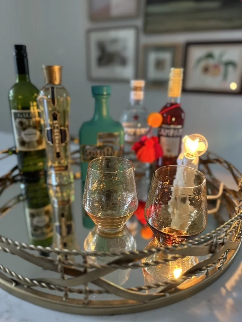 A decorative tray with assorted liquor bottles and two textured glasses on a reflective surface, featuring background artwork on a wall.