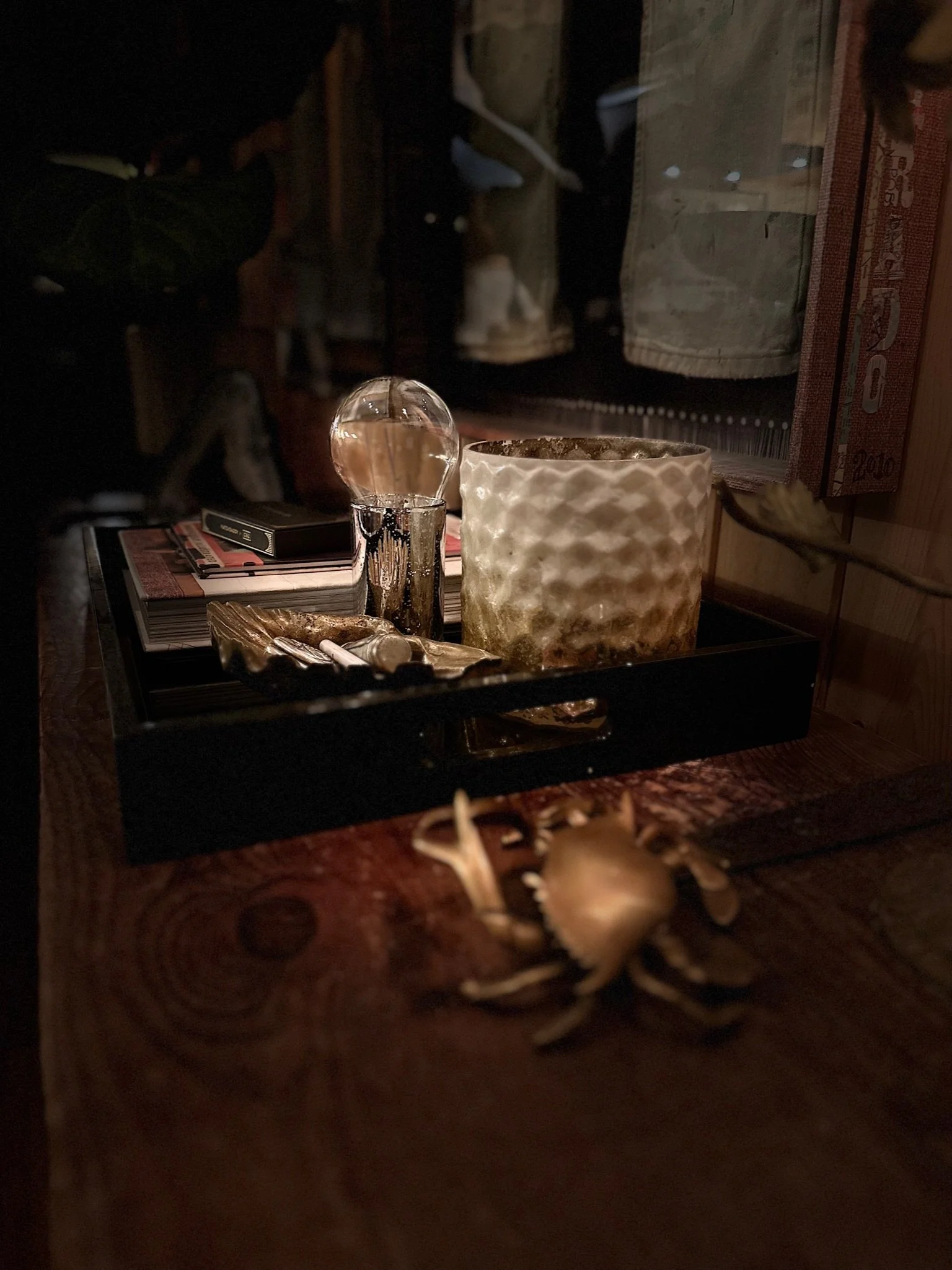 A dimly lit table with a decorative crab sculpture, a tray holding books, a gold-colored glass ornament, and a patterned candle holder. The background features fabric and books on a shelf.