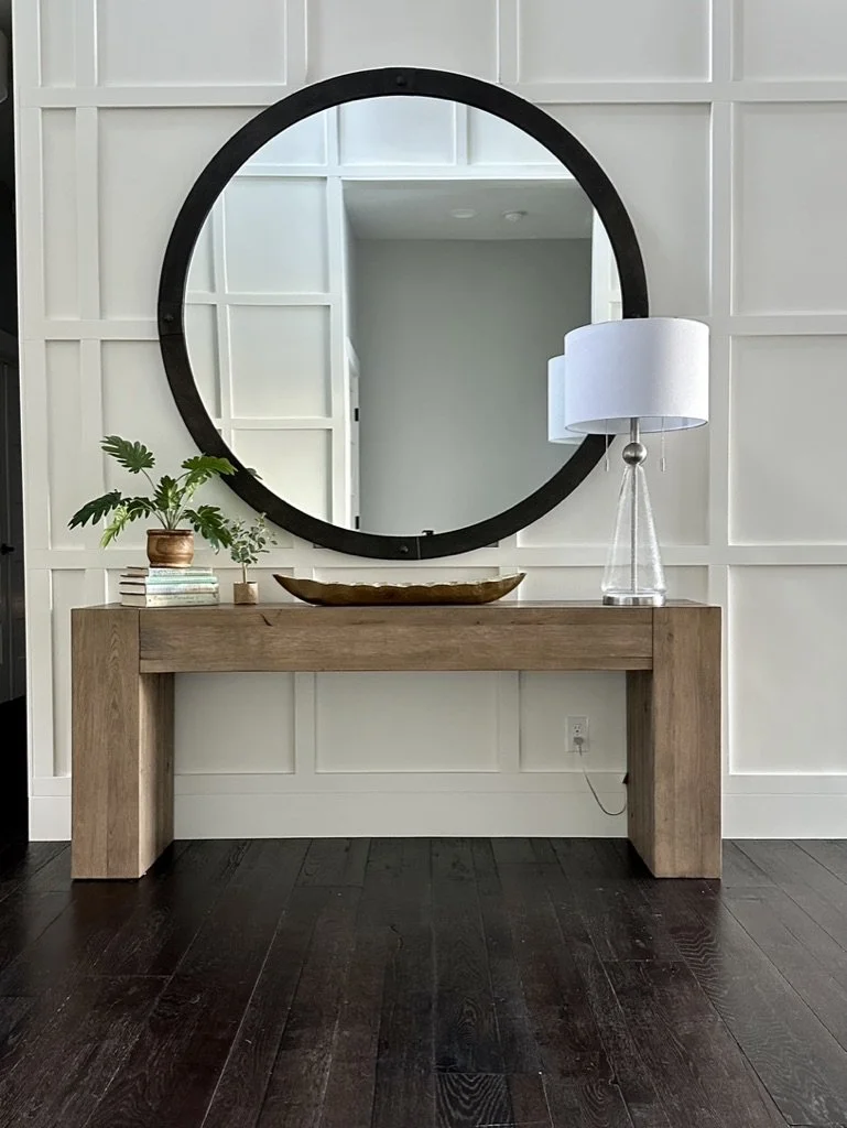 Modern entryway with large round mirror, wooden console table, lamp, decorative tray, houseplant, and books against paneled wall.