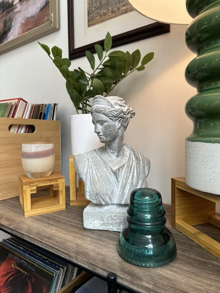 Bookshelf with decorative items including a bust sculpture, green glass insulator, potted plant, candle, and framed artworks on the wall.