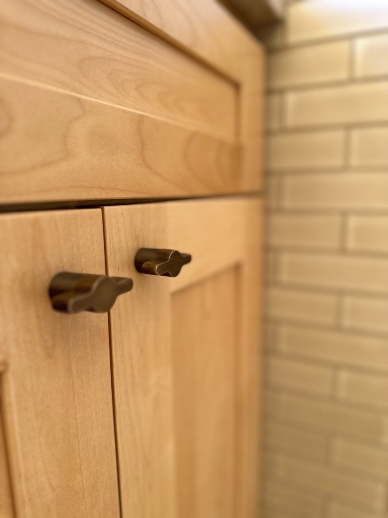 Close-up of light wood cabinet with brass handles next to beige tile wall.