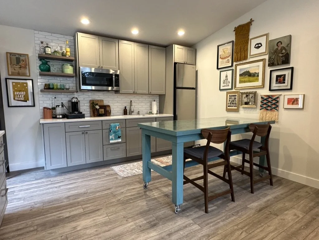 Modern kitchen with gray cabinets, stainless steel appliances, a teal table with two chairs, wooden floors, wall art, and shelves with various items.