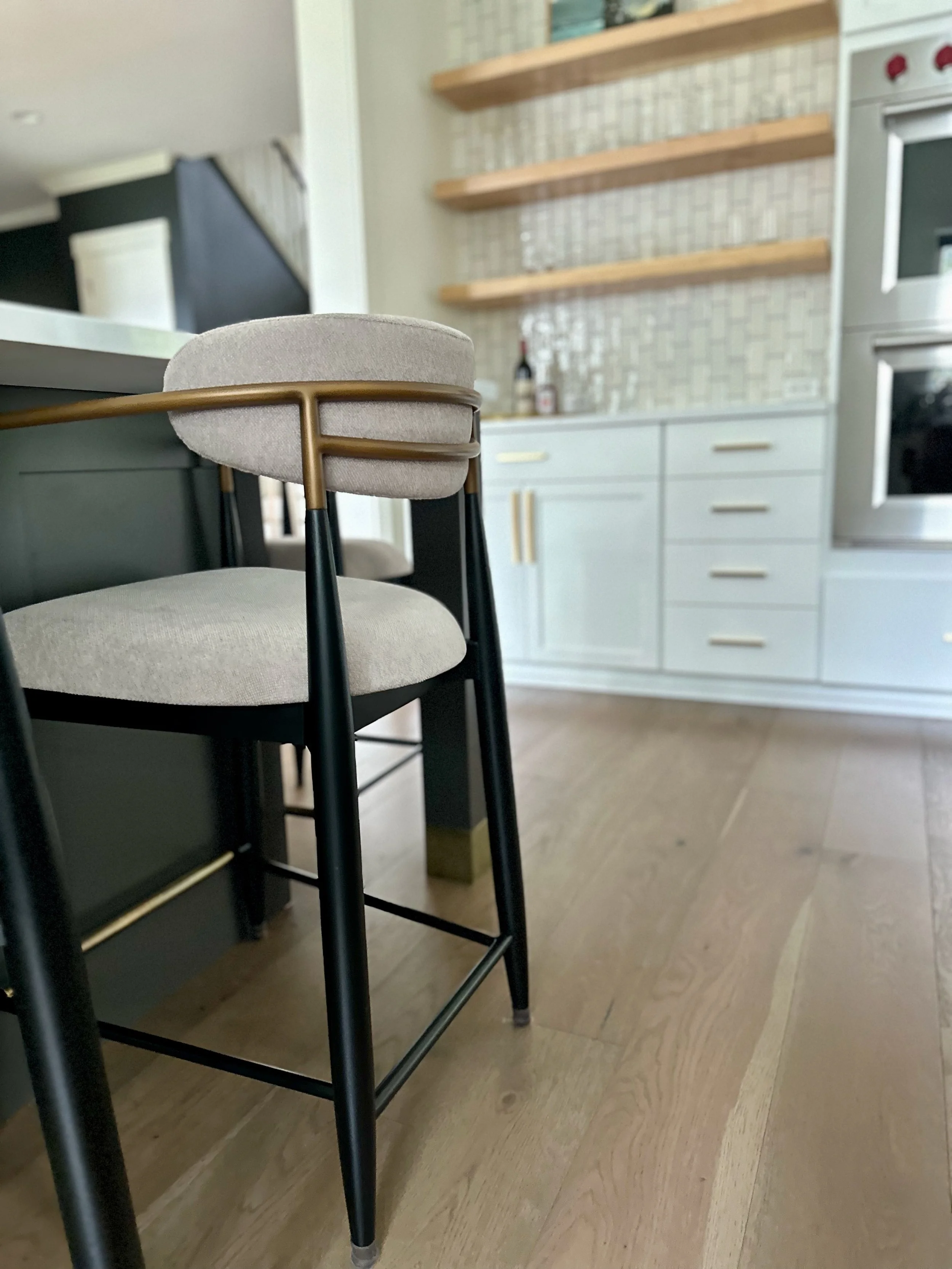 Modern kitchen interior with a black and gold bar stool placed near a kitchen island. Background features open wooden shelves, white cabinets, and stainless steel oven. Hardwood flooring is visible.