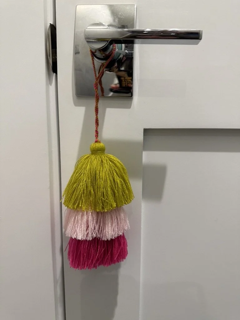 A modern silver door handle with a colorful tassel hanging from it. The tassel has three layers in green, pink, and magenta.