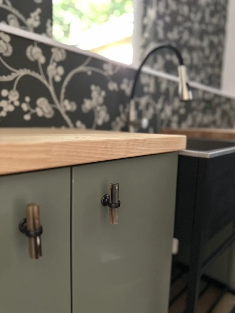Kitchen countertop with wood top and decorative black and white floral wallpaper, featuring a green cabinet with unique handles and a modern black faucet in the background.