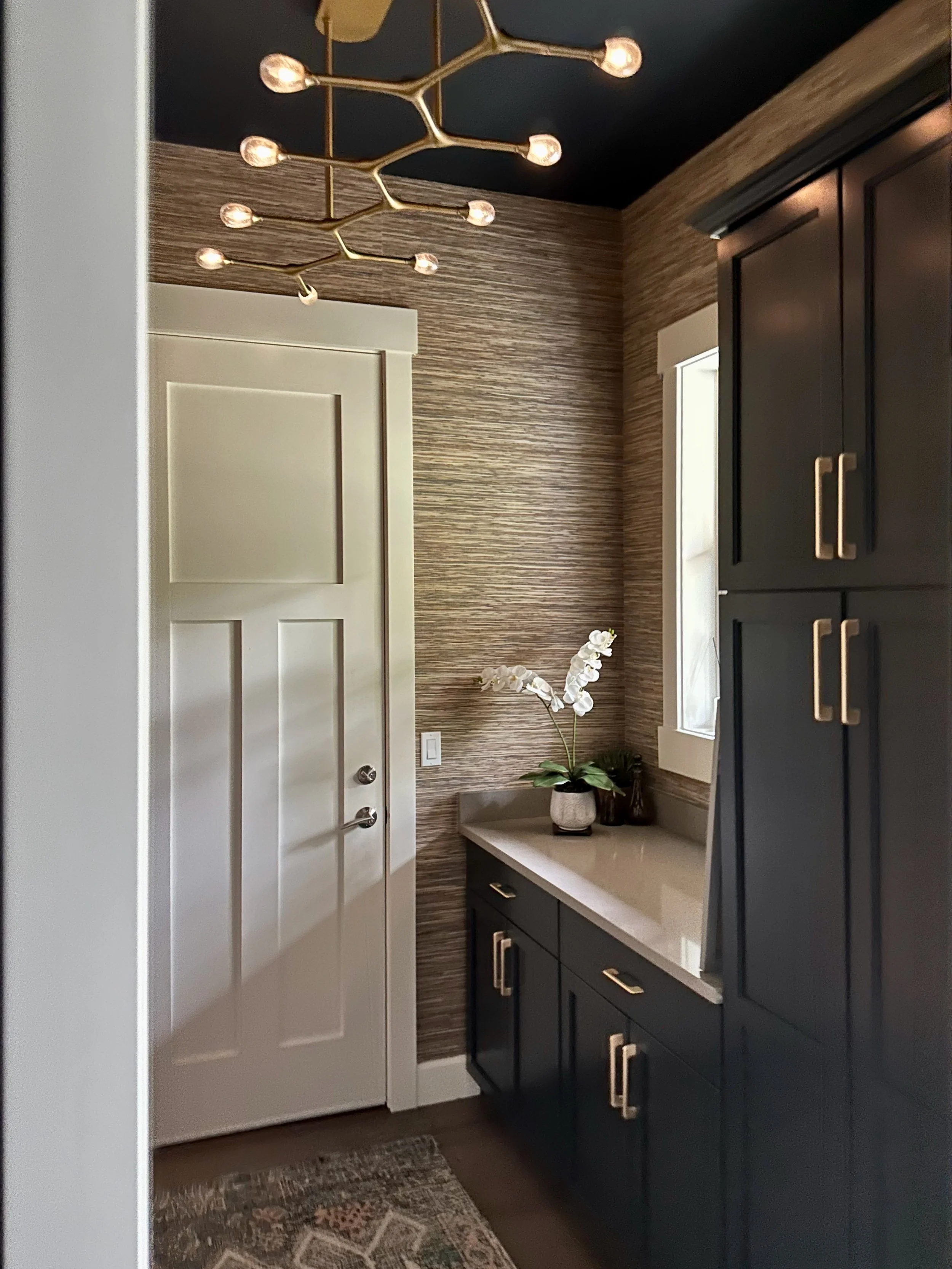 Modern mudroom with a dark blue cabinet featuring gold handles, a light-colored door, a unique gold branch chandelier, textured wallpaper, and a decorative plant on the countertop.