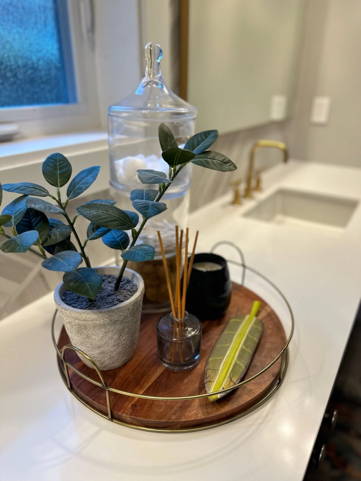 Bathroom countertop with decorative elements, including a potted plant, a glass jar with cotton balls, a black candle, reed diffuser, and a leaf-shaped item on a wooden tray.
