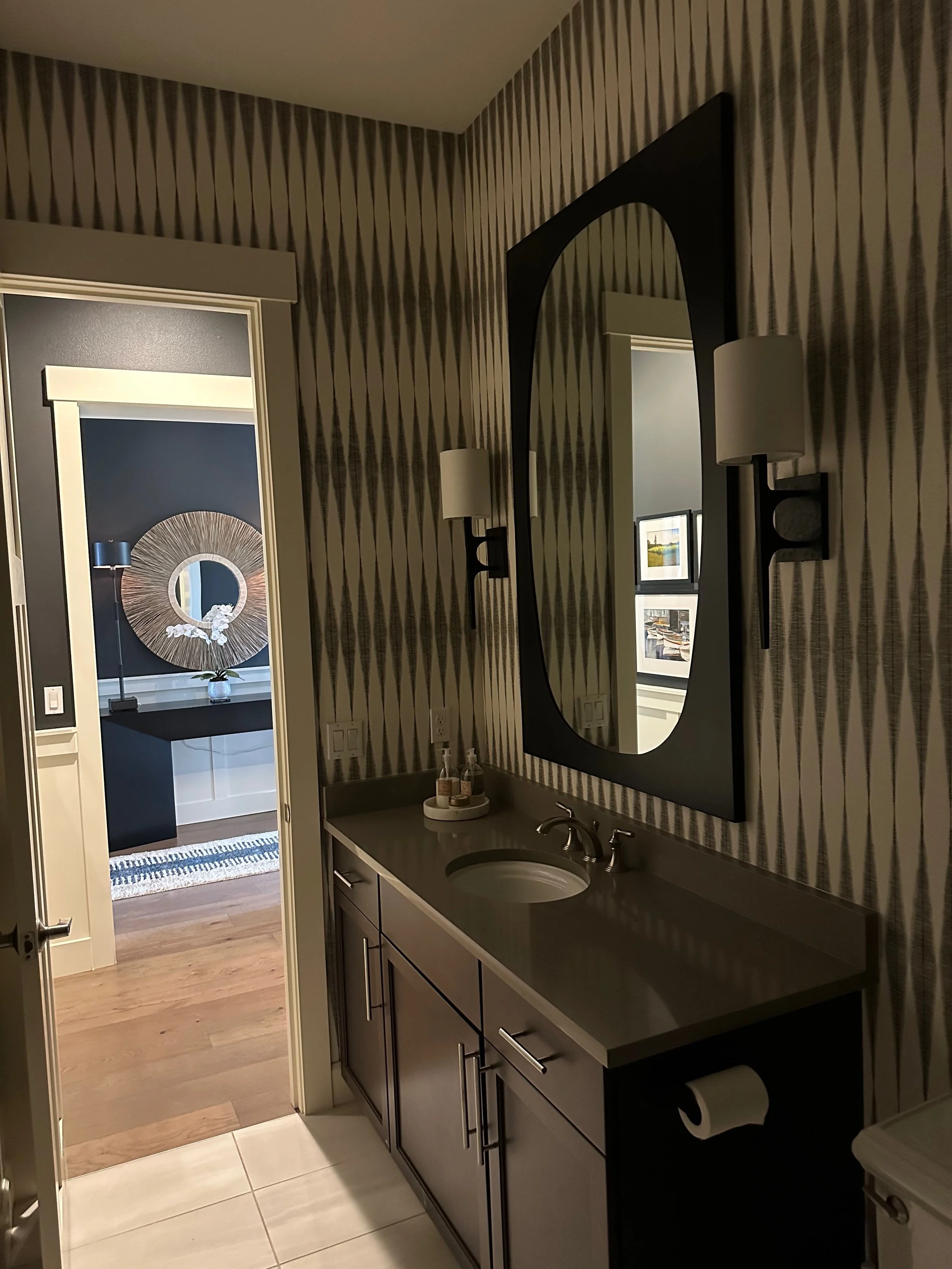 Modern bathroom with dark vanity, large mirror, geometric wallpaper, and a glimpse into a room with a round mirror and decorative items.