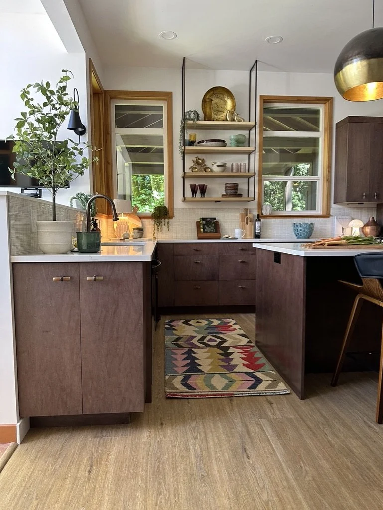 Modern kitchen with dark wood cabinets, white countertops, open shelving, and a colorful geometric rug. Indoor plants and decorative items are on shelves. Pendant light and two windows provide natural light.