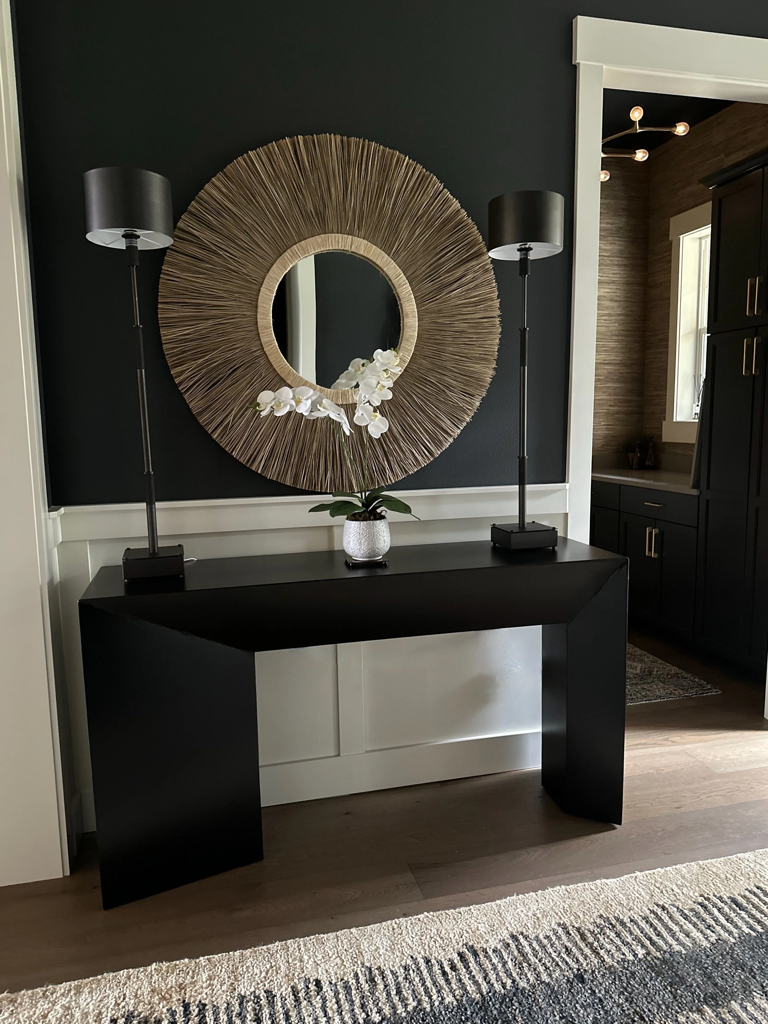 Modern entryway with a black console table, two black lamps, round mirror with straw frame, and potted white orchids on a textured wall.
