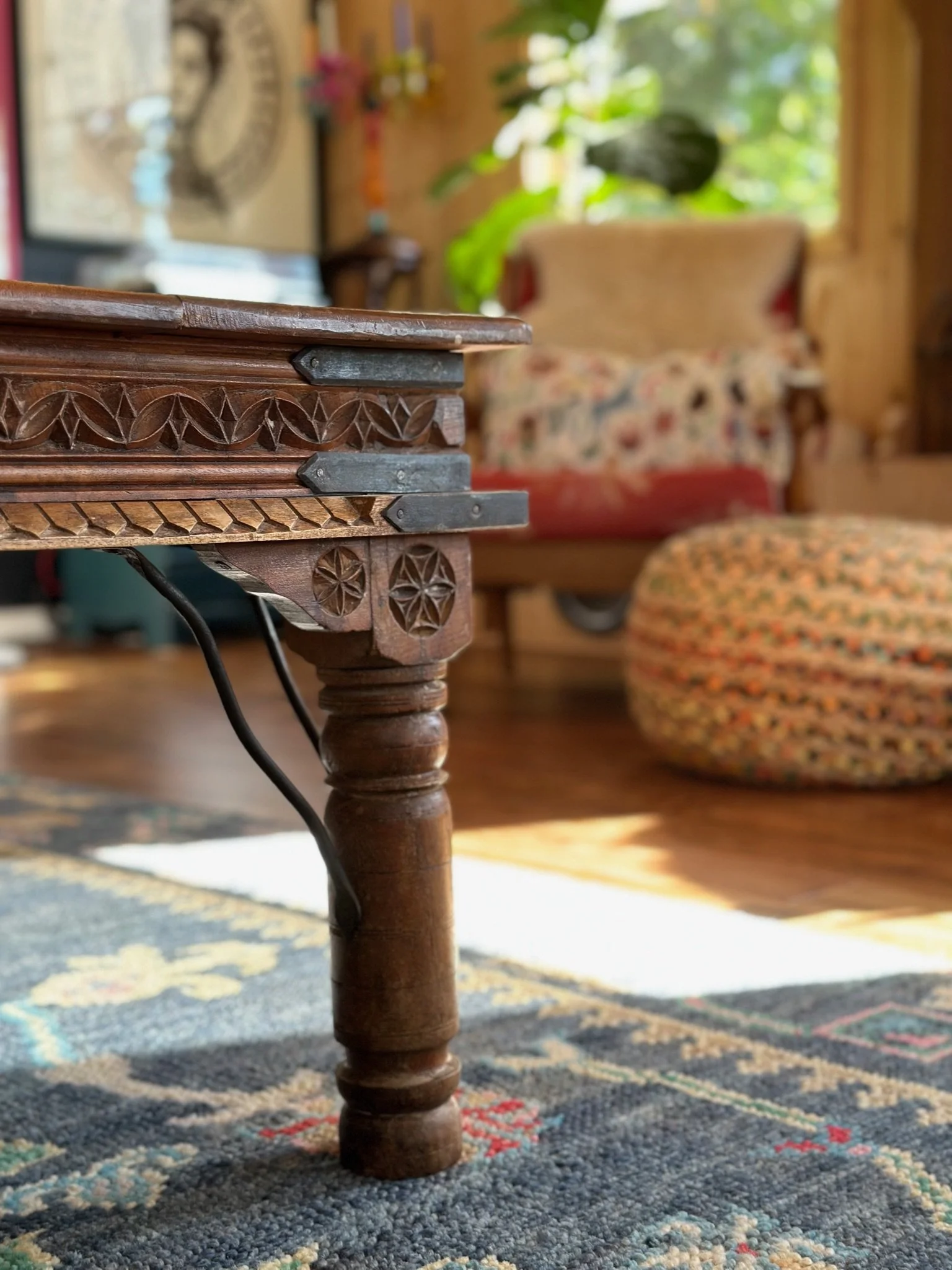 Close-up of a carved wooden table leg in a cozy, eclectic room with patterned rugs, an upholstered chair with a floral blanket, a braided pouf, and leafy plants in the background.