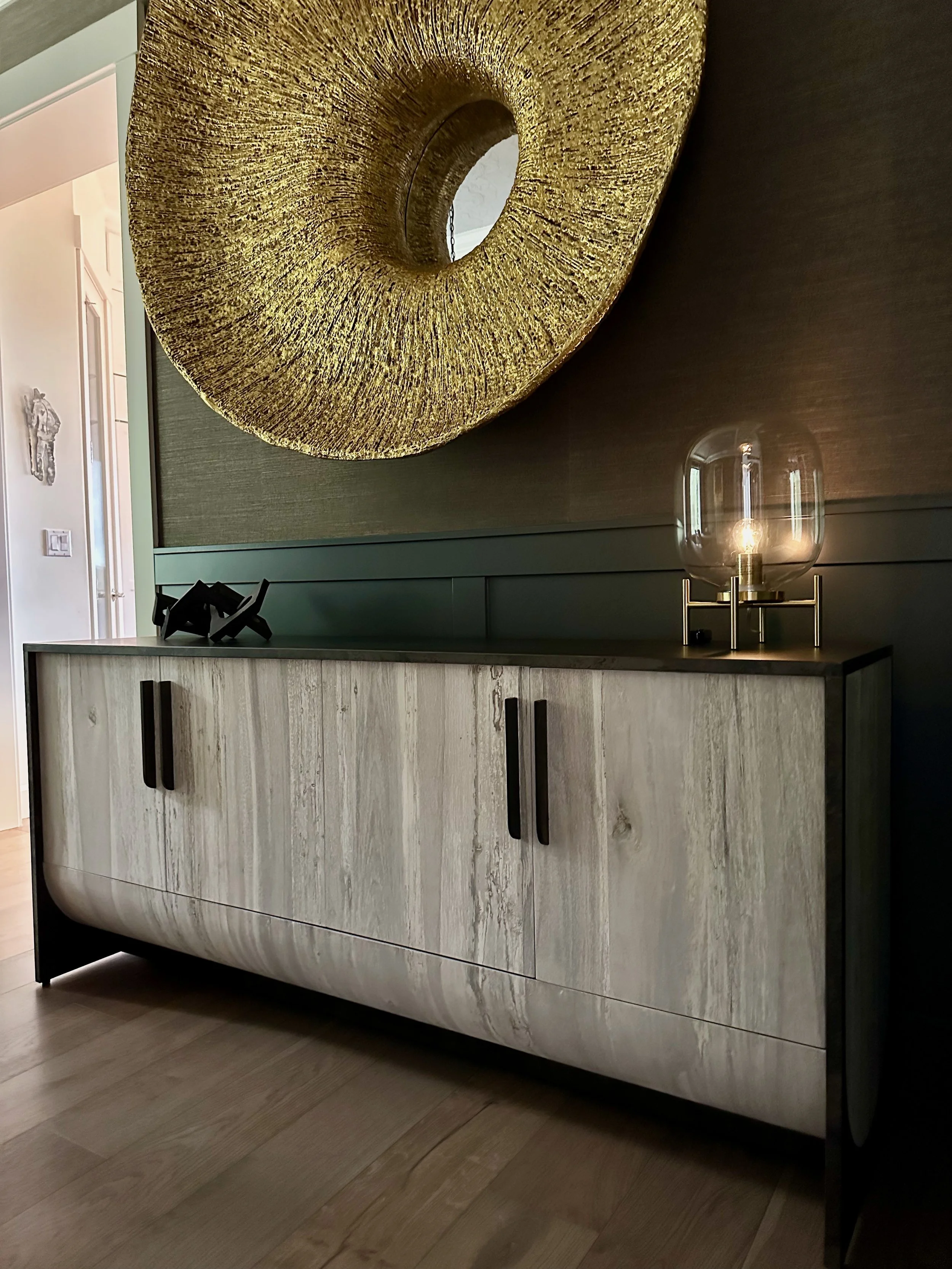 Modern gray sideboard with black handles placed against a dark wall, decorated with a large, textured gold circular wall art and a transparent lamp with an exposed bulb on top.