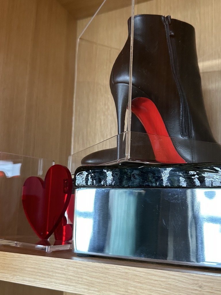 Black high-heeled boot with red sole in a display case next to red heart decor on a wooden shelf.