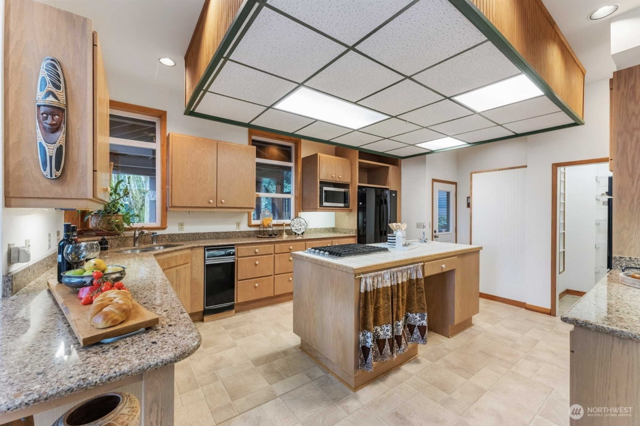 Modern kitchen with wooden cabinets, granite countertops, a central island with stovetop, and tile flooring. Features include a microwave, refrigerator, decorative mask, and assorted fresh fruit on a cutting board.