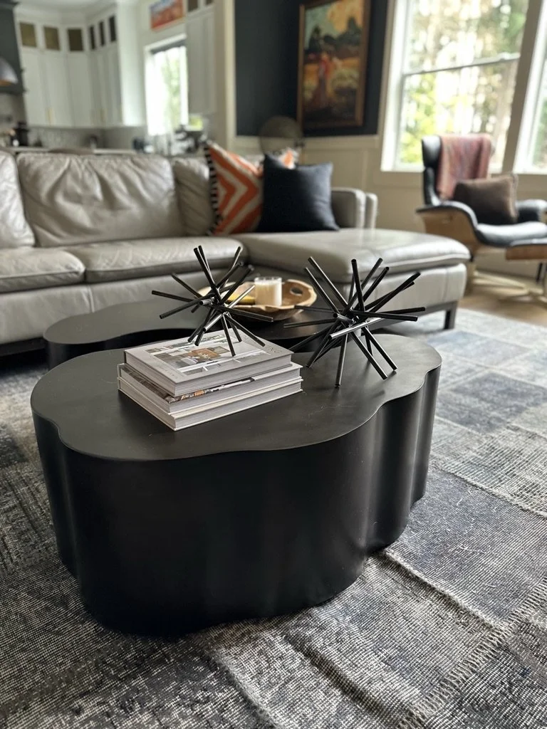 Modern living room with a black, organic-shaped coffee table featuring geometric sculptures, books, and a candle. Grey sofa with orange and black accent pillows in the background, next to a window and a framed artwork.