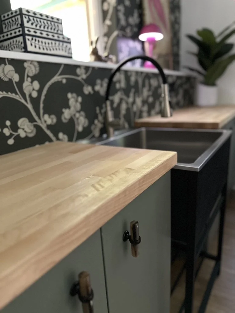 Modern kitchen with wooden countertops, a stainless steel sink, floral wallpaper, decorative boxes, a lamp, and a potted plant.