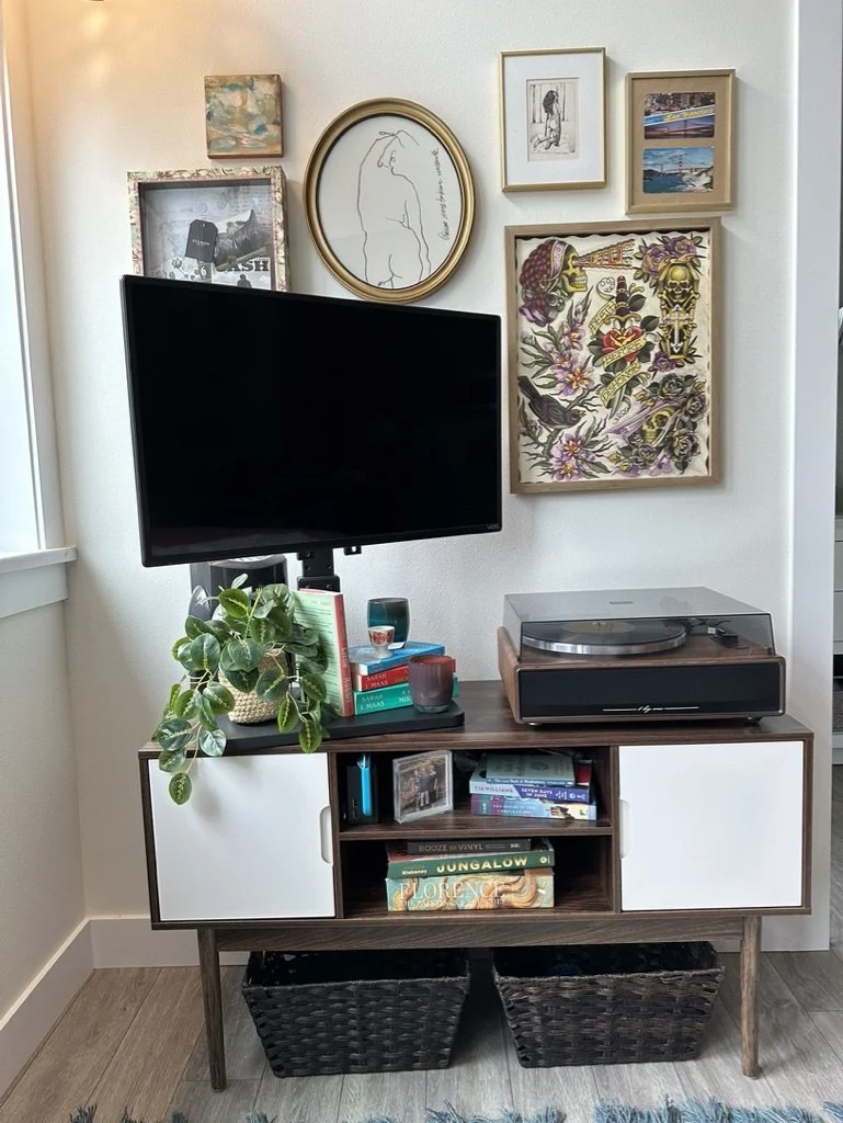 Living room setup with a television mounted on a wall above a mid-century style cabinet. The cabinet holds a record player, books, and decorative items. Wall art and framed pictures are displayed above. A potted plant is placed on the cabinet, and tw